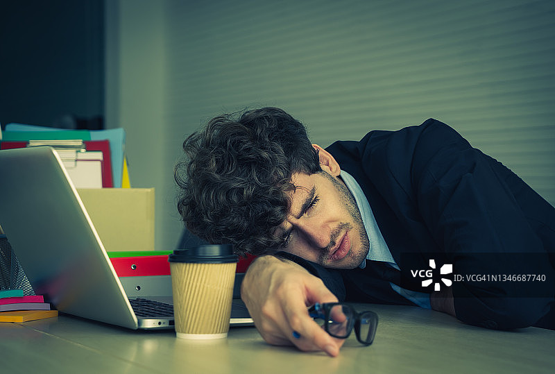 疲惫的商人在他的办公桌上打盹在办公室的笔记本电脑和咖啡的办公室过度工作的概念。图片素材