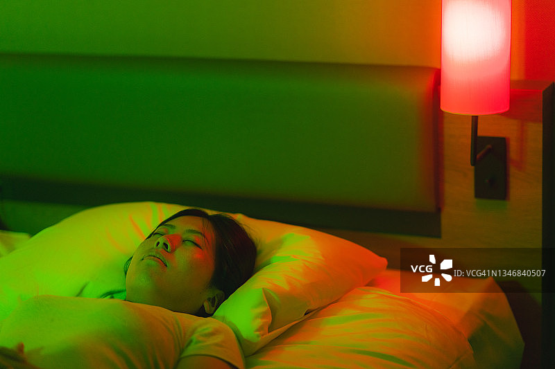年轻女子被红色和绿色的霓虹灯照亮，躺在家里卧室的床上休息图片素材