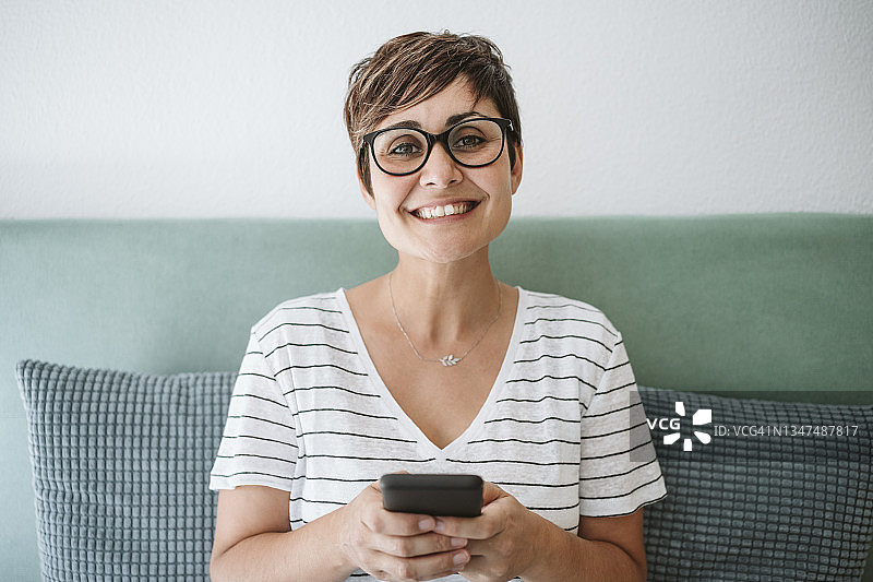 快乐的中年女性戴着眼镜，在家拿着智能手机图片素材