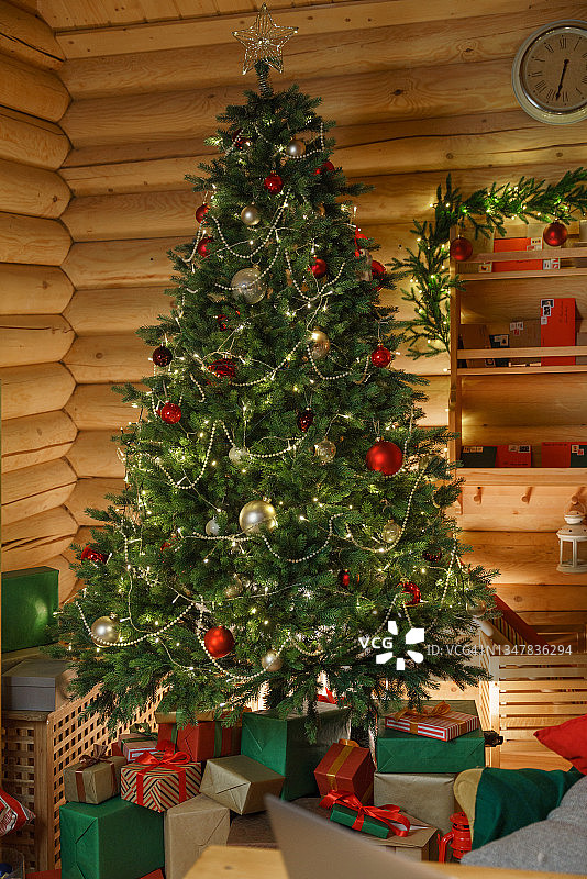 一棵装饰精美的圣诞树，下面有玩具、圣诞灯和许多礼物，我们看到它在温暖的电灯照明下的全长图片素材