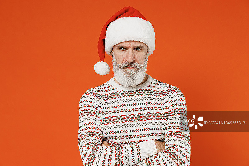 老的严厉的阴沉的忧郁的疲倦的疲倦的严厉的皱眉的胡子圣诞老人男人50多岁穿着圣诞帽毛衣摆姿势看相机手交叉孤立在普通的橙色背景摄影棚。图片素材