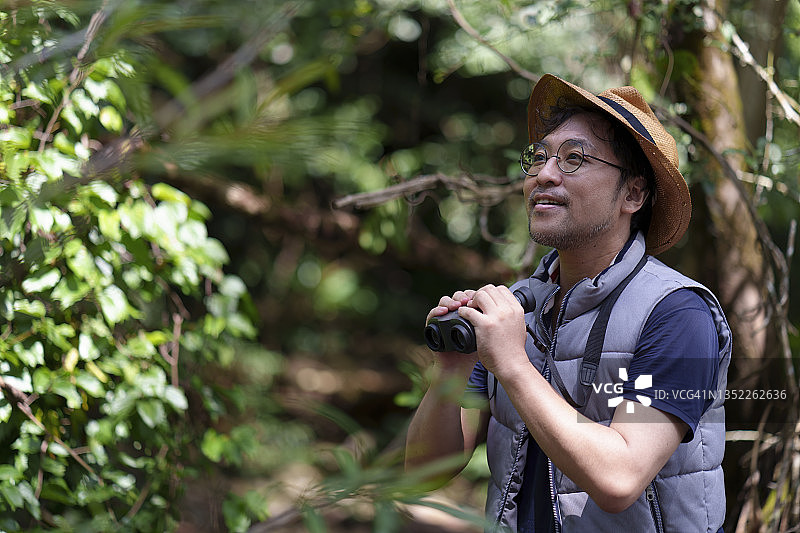 一名日本男子手持双筒望远镜站在森林露营地，环顾四周。图片素材