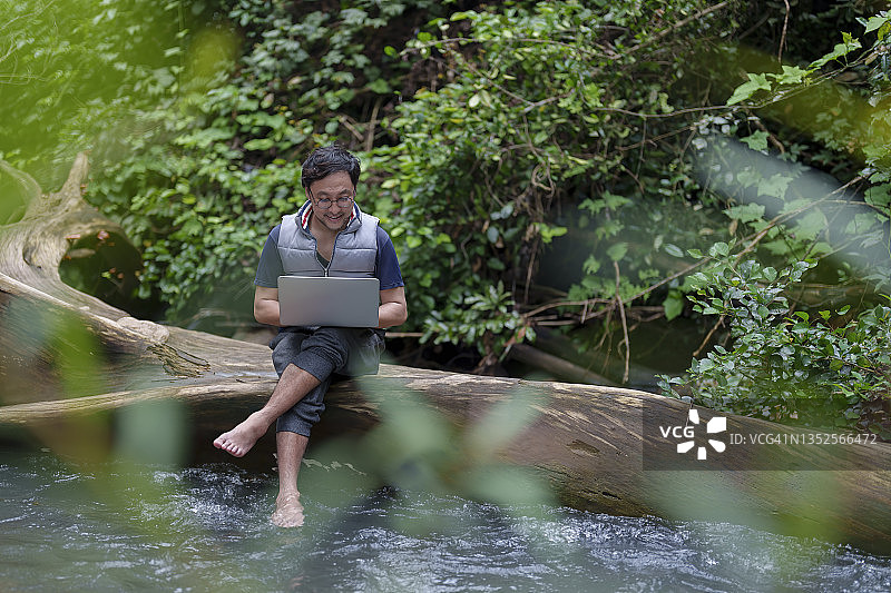 一名使用笔记本电脑的日本男子在工作和度假期间喜欢在自然的溪流中工作。图片素材