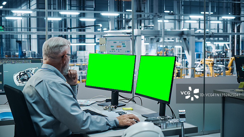 汽车工厂监控办公室:男首席汽车工程师坐在办公桌前工作的绿色屏幕色度键电脑。自动化机器人手臂装配线制造车辆图片素材