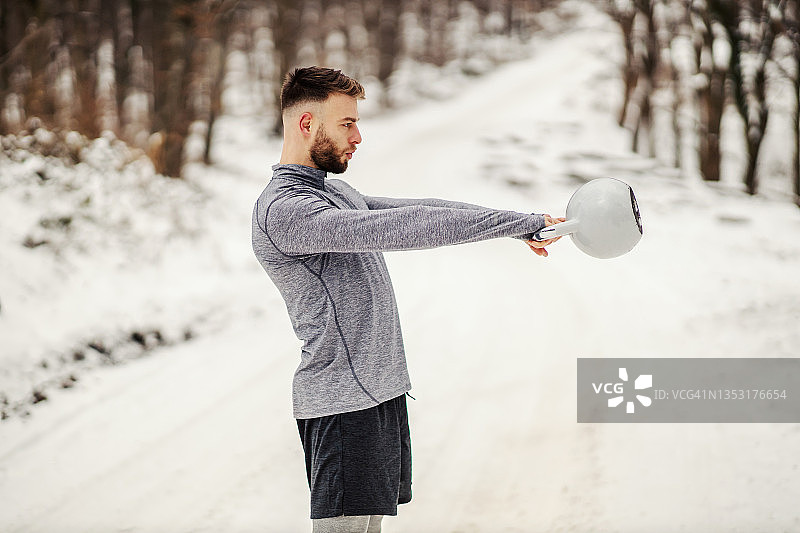 健美运动员站在森林的雪道上，摇摆着冬天的壶铃。健身运动，冬季健身，户外健身图片素材
