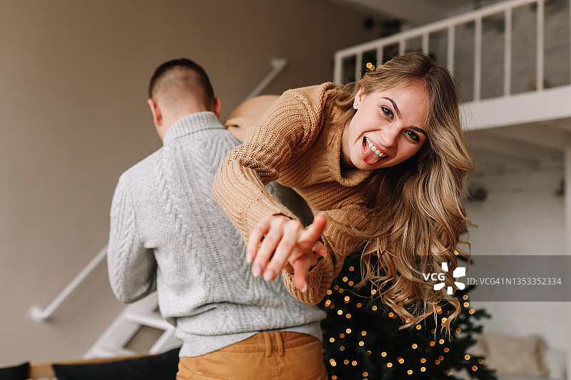 在圣诞节周末，一对穿着毛衣的幸福的情侣在舒适的房子里装饰着圣诞树的房间里开怀大笑，在冬天的室内过新年图片素材