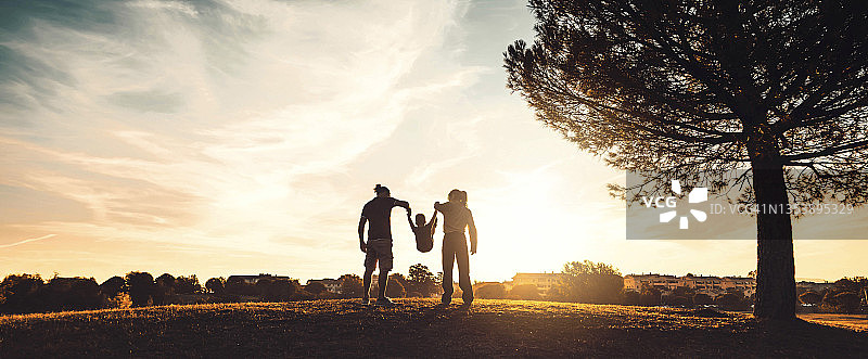夕阳下在草地上散步的幸福家庭的剪影——母亲、父亲和儿子在户外一起享受时光——家庭、爱、心理健康和快乐的生活理念图片素材