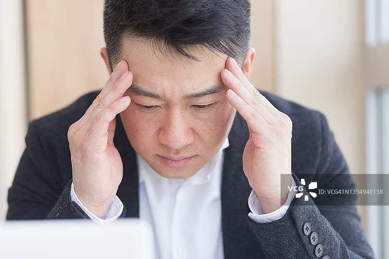 年轻的亚洲男子，办公室职员坐在工作场所握着手按摩额头和头部与严重的疼痛。工作时头痛的男性。近距离接触患者的脸是超负荷的疾病。特写镜头图片素材