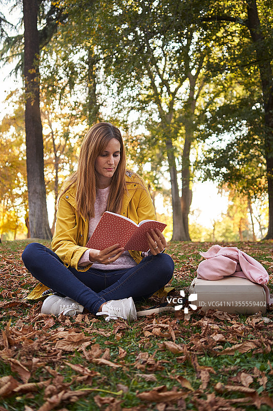 穿着便服和黄大衣的年轻女子在树木环绕的公园里坐着看书。24、闲暇观，自然观，读书观。图片素材