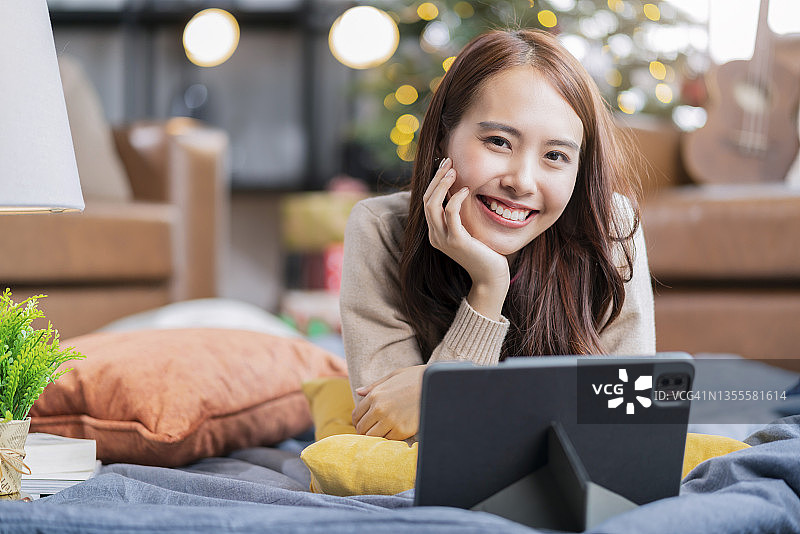 年轻的有吸引力的亚洲女性呆在家里使用平板电脑技术设备视频打电话给她的朋友或家人的问候和圣诞节庆祝在客厅的沙发附近的节日概念孤立图片素材