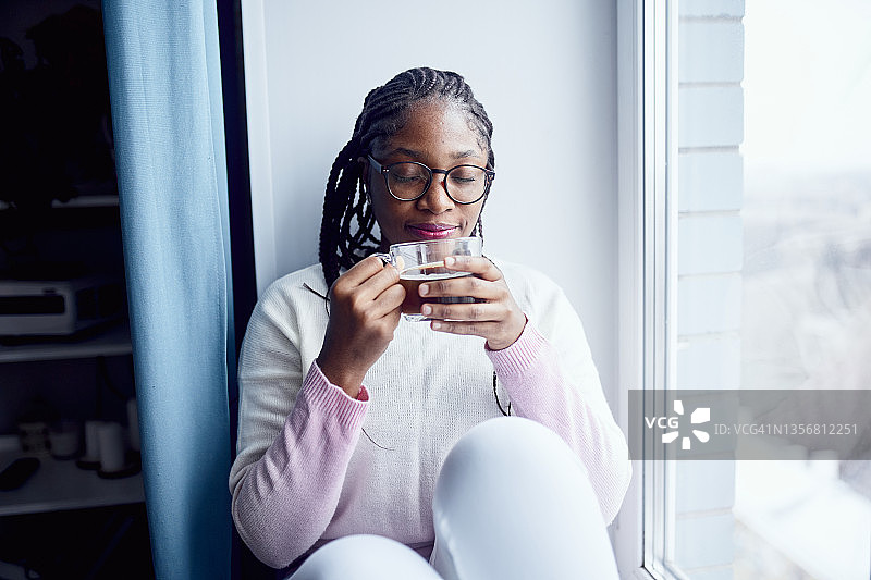 一位年轻女子坐在窗边，闭着眼睛享受着喝咖啡的乐趣图片素材