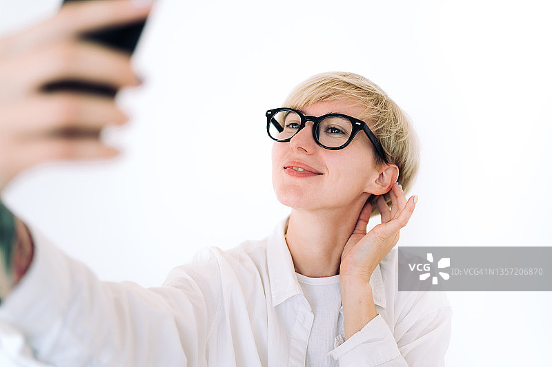 金发短发、戴眼镜的年轻女子正在用智能手机自拍。她穿着白色的中性休闲服装。性别中立概念图片素材