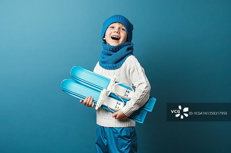 手里拿着迷你滑雪板的有趣小孩。冬季运动和休闲概念，工作室拍摄浅蓝色背景。图片素材