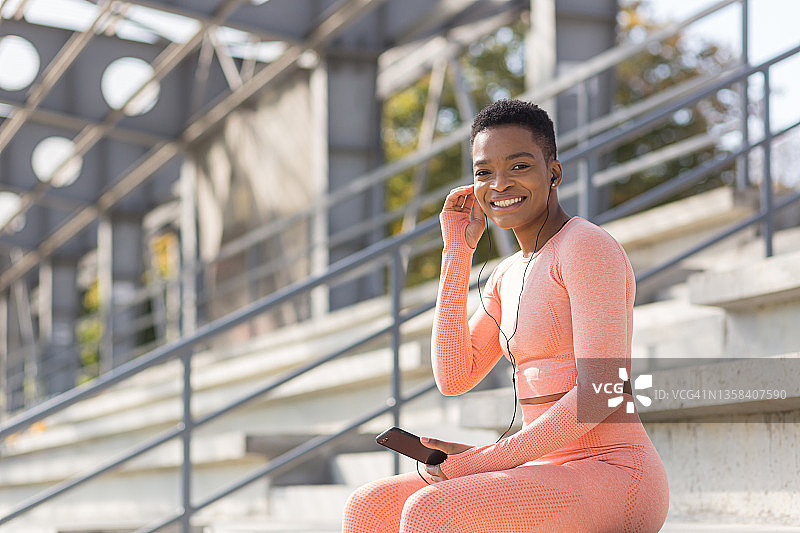 美丽和成功的女运动员微笑着听有声书从移动电话在体育馆附近晨练和慢跑，非洲裔美国妇女与移动电话图片素材