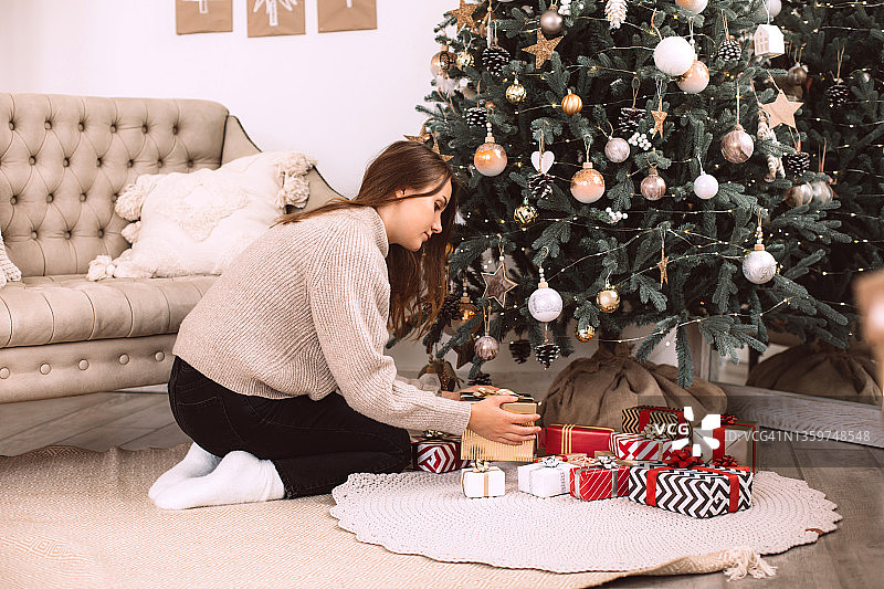 在装饰好的圣诞树下，一名女子正拿着金色的礼品盒，在编织地毯上摆放着许多不同的礼品盒。新年庆祝理念。前视图图片素材