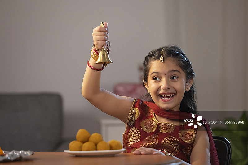 一个穿着传统服装的小女孩高兴地摇着铃铛，桌上放着一盘土豆。图片素材