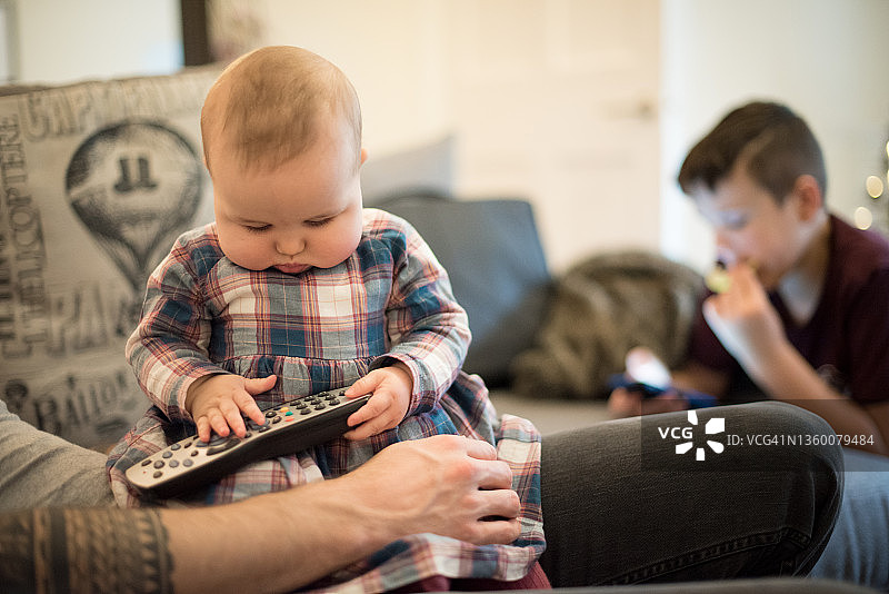 在圣诞节期间，胖乎乎的宝宝在爸爸的腿上玩电视遥控器，而她十几岁的弟弟正在查看他的智能手机图片素材