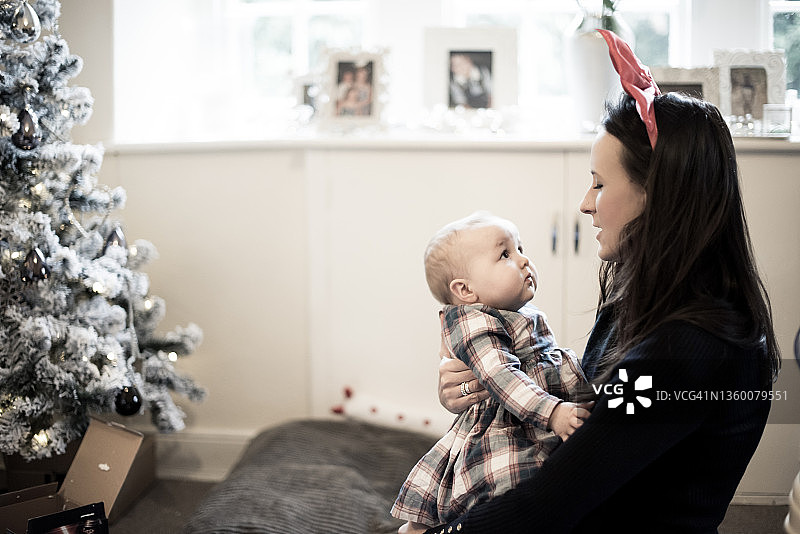 年轻的母亲戴着红色的圣诞蝴蝶结头带，抱着她的孩子在她的手臂和她说话旁边的白色圣诞树图片素材