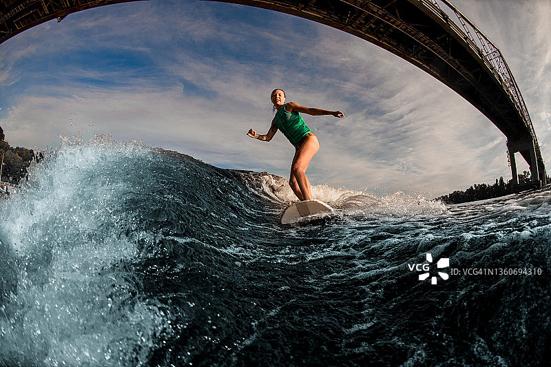 穿着绿松石马甲的女子在溅起的波浪冲击着桥面的冲浪板上冲浪。图片素材