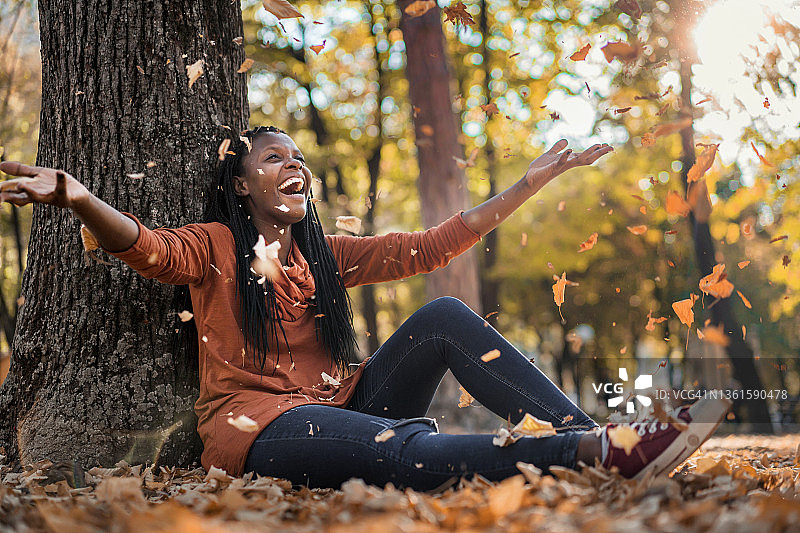 微笑的黑人妇女坐在一棵树前的地上，扔着秋叶图片素材