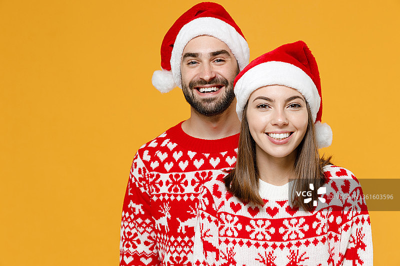 微笑的年轻圣诞老人夫妇，两个朋友，男人，20多岁的女人穿着红色毛衣，圣诞帽看着相机孤立在黄色背景工作室的肖像。祝新年庆佳节快乐理念。图片素材