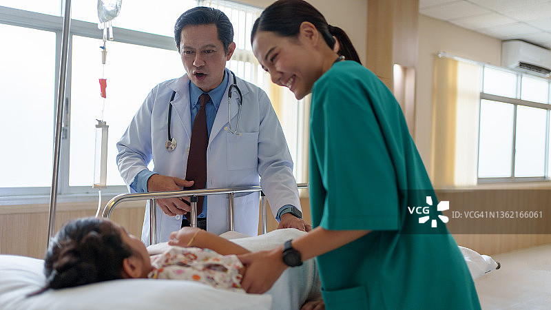 亚洲男医生安慰和谈话的女孩病人在医院病房医疗访问期间与女护士站在背景中。图片素材