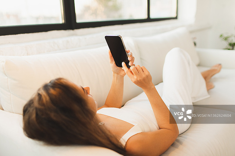 无聊的女人躺在沙发上玩手机。把时间浪费在网络上。拖延症与设备图片素材