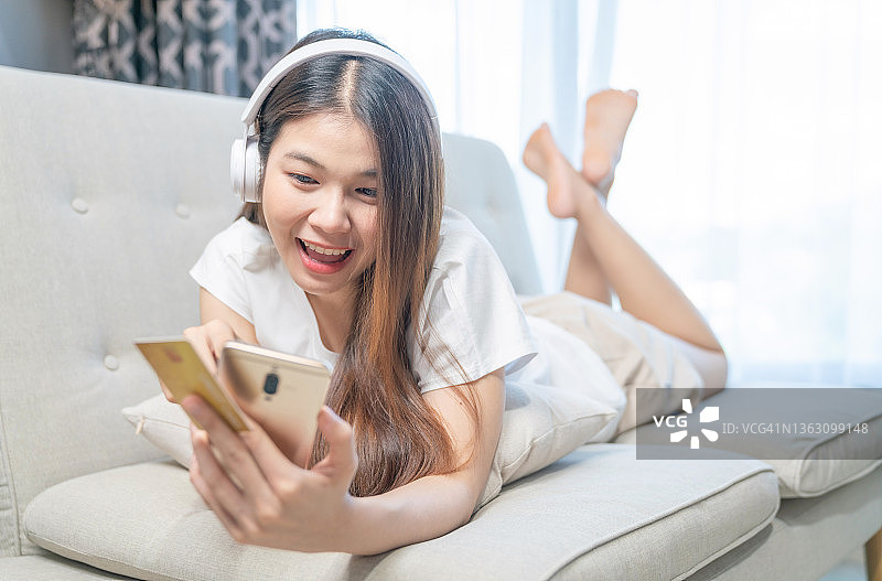 年轻微笑的亚洲妇女使用智能手机购买网上购物信用卡，而躺在沙发时，放松在客厅在家里。生活方式拉丁和西班牙裔妇女在房子的概念。图片素材
