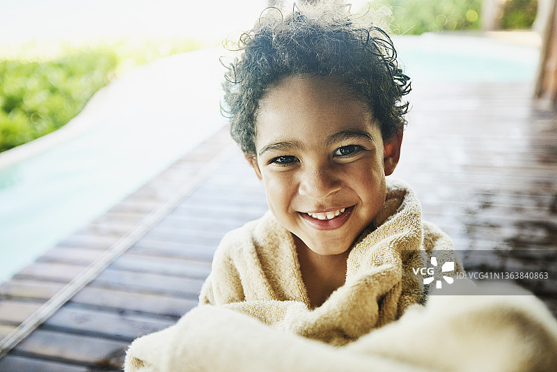 中近镜头的照片，微笑的小男孩裹在毛巾后，在度假游泳池游泳图片素材