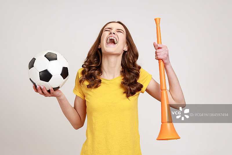 穿着黄色t恤的少女兴奋地拿着喇叭和黑白相间的足球，庆祝自己最喜欢的足球队在冠军比赛中获胜。图片素材