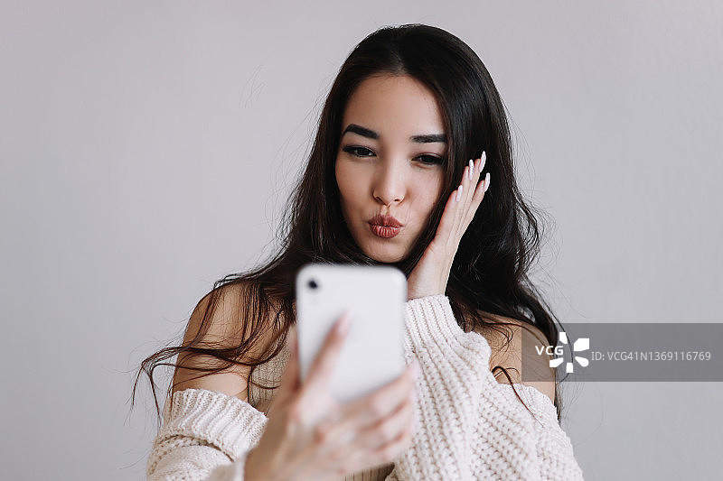 一名年轻的亚洲千禧一代女性拍了一张自拍，并用无线技术在手机上交流。一个愉快微笑的女孩在一个白色孤立的背景网上视频聊天的大肖像图片素材