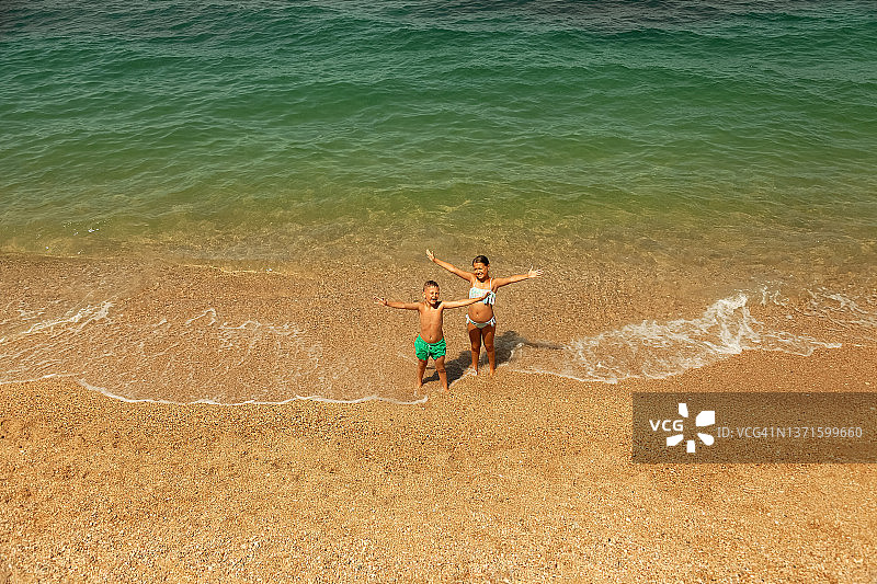 在一个夏日的沙滩上，一对穿着泳衣的男孩和女孩站在湛蓝的海边，两人都举起手来打招呼图片素材