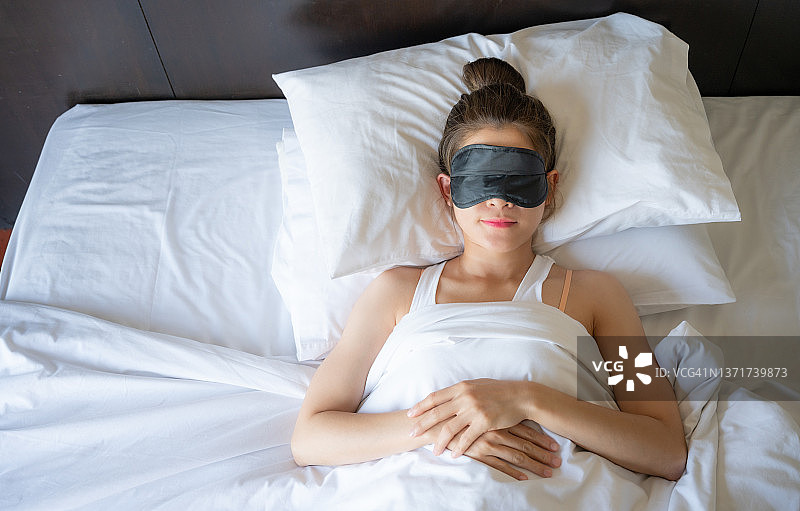 高角度的年轻亚洲妇女戴着睡眠面罩在床上睡觉。图片素材