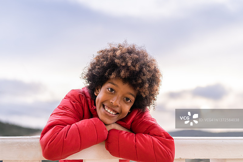 一个穿着红夹克，面带微笑的黑人男孩倚在栏杆上图片素材