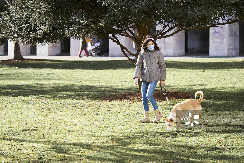 一个冬天的早晨，女孩穿着夹克和御寒靴在公园遛狗，戴着防护口罩，感染了新冠肺炎。图片素材