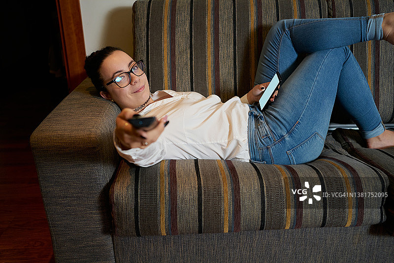 年轻女子一边微笑一边将电视遥控器对准摄像机。一个女人躺在客厅沙发上玩手机。图片素材