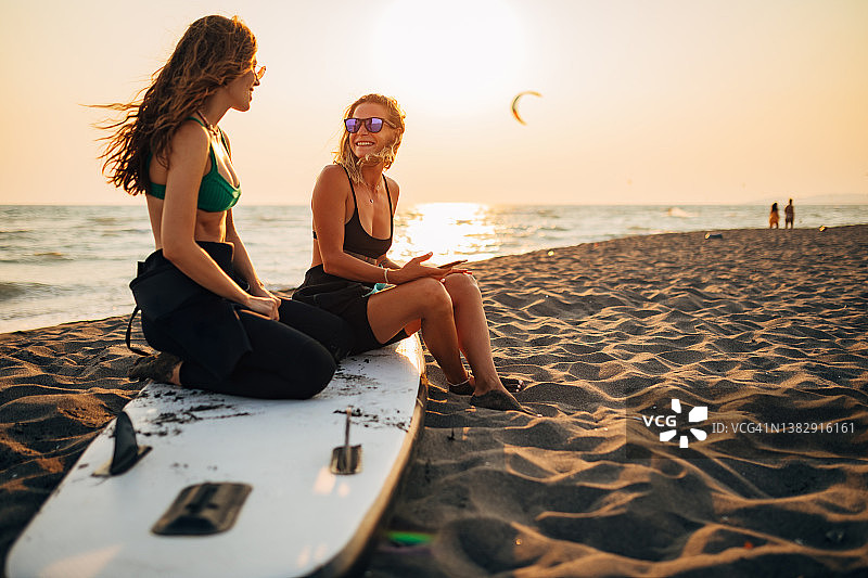 两个美丽的冲浪者坐在沙滩上，在SUP板上聊天，享受一起度过的自由时间。图片素材