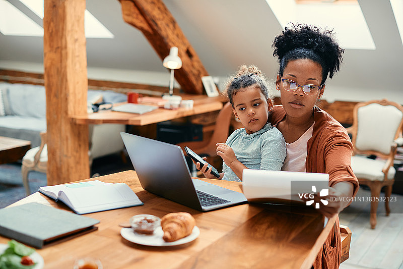 非裔美国职业母亲使用笔记本电脑和分析文件，而女儿坐在她的腿上在家里。图片素材