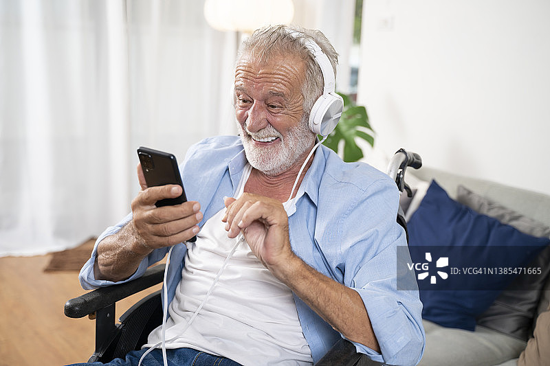 快乐的残疾老人在轮椅上用智能手机和耳机听他最喜欢的音乐在家里的客厅里图片素材