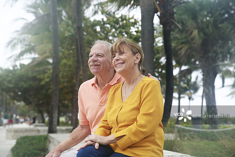 在多云的天气，一对成熟的白人夫妇穿着色彩鲜艳的上衣，愉快地坐在海边的长椅上。图片素材
