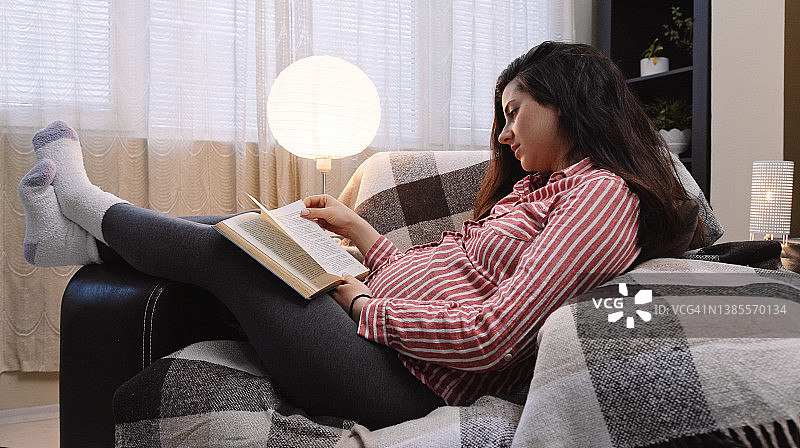 一位孕妇正跷着脚坐在舒适的扶手椅上看书。图片素材