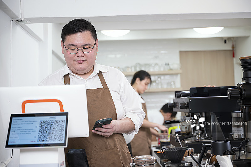 在收银台，一名亚裔男性咖啡店老板正在查看顾客的订单，而女咖啡师正在按顾客的订单制作咖啡。图片素材