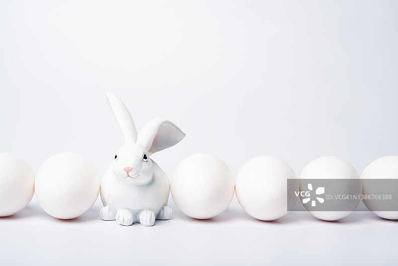 一排白色的鸡蛋和一个玩具兔子在白色的背景上。复活节假期的概念。单色背景。图片素材