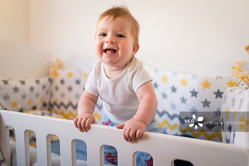 面带微笑，快乐的白人男婴站在婴儿床上图片素材