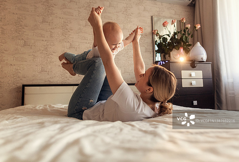 一个年轻的妈妈微笑着躺在一张舒适的床上，把她的小女儿抱在腿上。一个快乐的小女孩在空中飞翔，时刻温柔地和孩子玩耍。图片素材