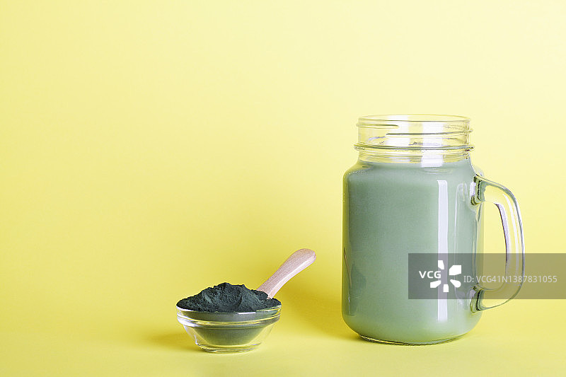 新鲜的绿色冰沙，玻璃瓶和螺旋藻粉，勺子在黄色的背景。有益的习惯，自我照顾和健康的生活方式。本空间图片素材