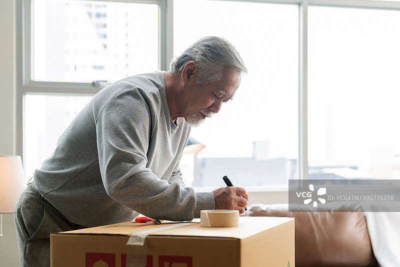 水平中景拍摄成年老年亚洲人男性白胡子写东西整理卡片盒准备搬到新房子打包东西，并把它们粘在客厅，搬家的想法概念图片素材