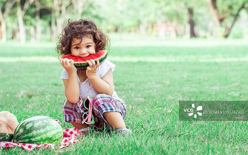 白种可爱的小女孩坐着，在户外的绿色公园里玩野餐，拿着，吃着，咬着水果，吃着新鲜的西瓜。教育，自然，食品概念。图片素材