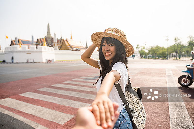 在曼谷旅行时，一位年轻的亚洲女性拉着男友的手穿过人行横道图片素材