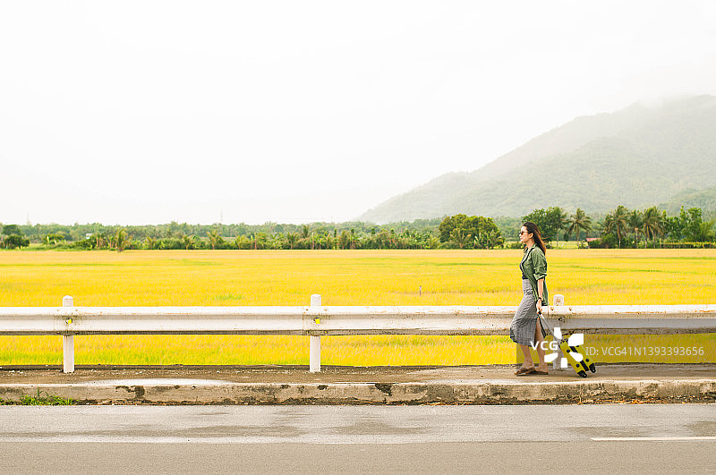 一位年轻的东南亚女性提着行李走在黄色稻田旁的道路上图片素材